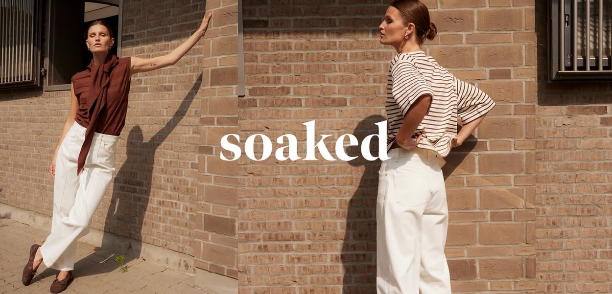 Zwei Frauen stehen vor einer sonnenbeschienenen Backsteinmauer und tragen weiße Hosen; eine trägt ein braunes ärmelloses Oberteil, die andere ein gestreiftes kurzärmeliges Hemd.