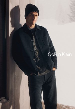 Giacca di lana scura sopra un maglione grigio testurizzato, abbinata a pantaloni neri, con sfondo di neve coperta e parete di legno.