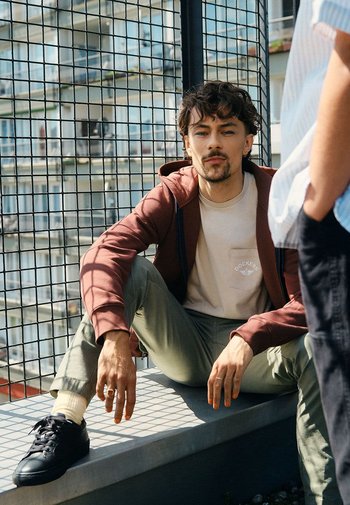 Le modèle est assis sur un rebord, portant un sweat à capuche bordeaux, un t-shirt crème avec une poche, un pantalon vert olive et des baskets noires avec des semelles blanches.