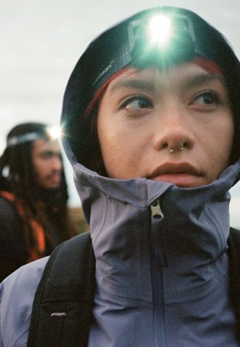 Frontala cu LED strălucitor, purtată pe o jachetă cu glugă întunecată. Are un fermoar și o textură moale. Fundalul include o persoană cu frontala.