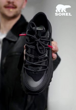 Personne tenant une basket noire Sorel avec des lacets à motifs, logo visible sur la languette de la chaussure et dans le coin supérieur droit de l'image.