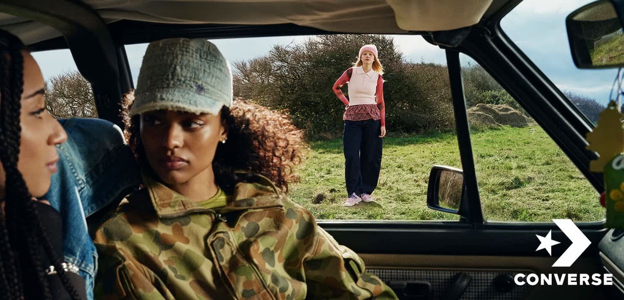 Dos mujeres sentadas en un coche, una con chaqueta y gorra de camuflaje, mirando a una mujer de pie afuera sobre el césped, vestida con ropa rosa y roja.