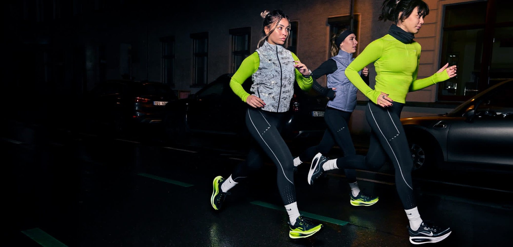 Drei Frauen laufen nachts in einer Stadt, tragen leuchtend grüne Sportoberteile, schwarze Leggings mit reflektierenden Akzenten und gemusterte Westen.