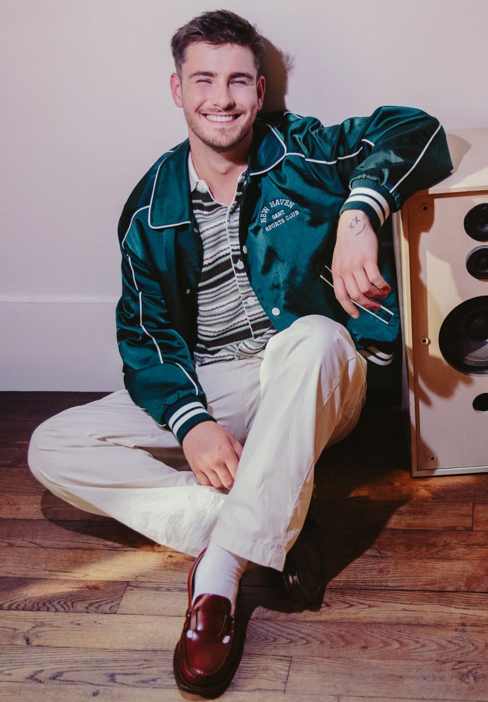 Groene satijnen bomberjack met witte bies, gemusterde shirt, crèmekleurige broek en bruine loafers. Luidspreker naast persoon die op de houten vloer zit.