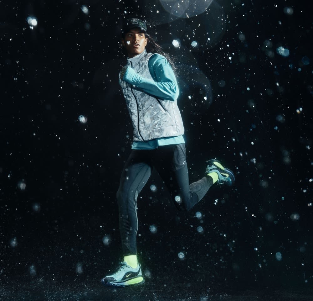Coureur portant un gilet réfléchissant argenté, un haut à manches longues bleu clair, un legging noir et des chaussettes vert fluo. Les chaussures de course arborent des accents noirs et verts.