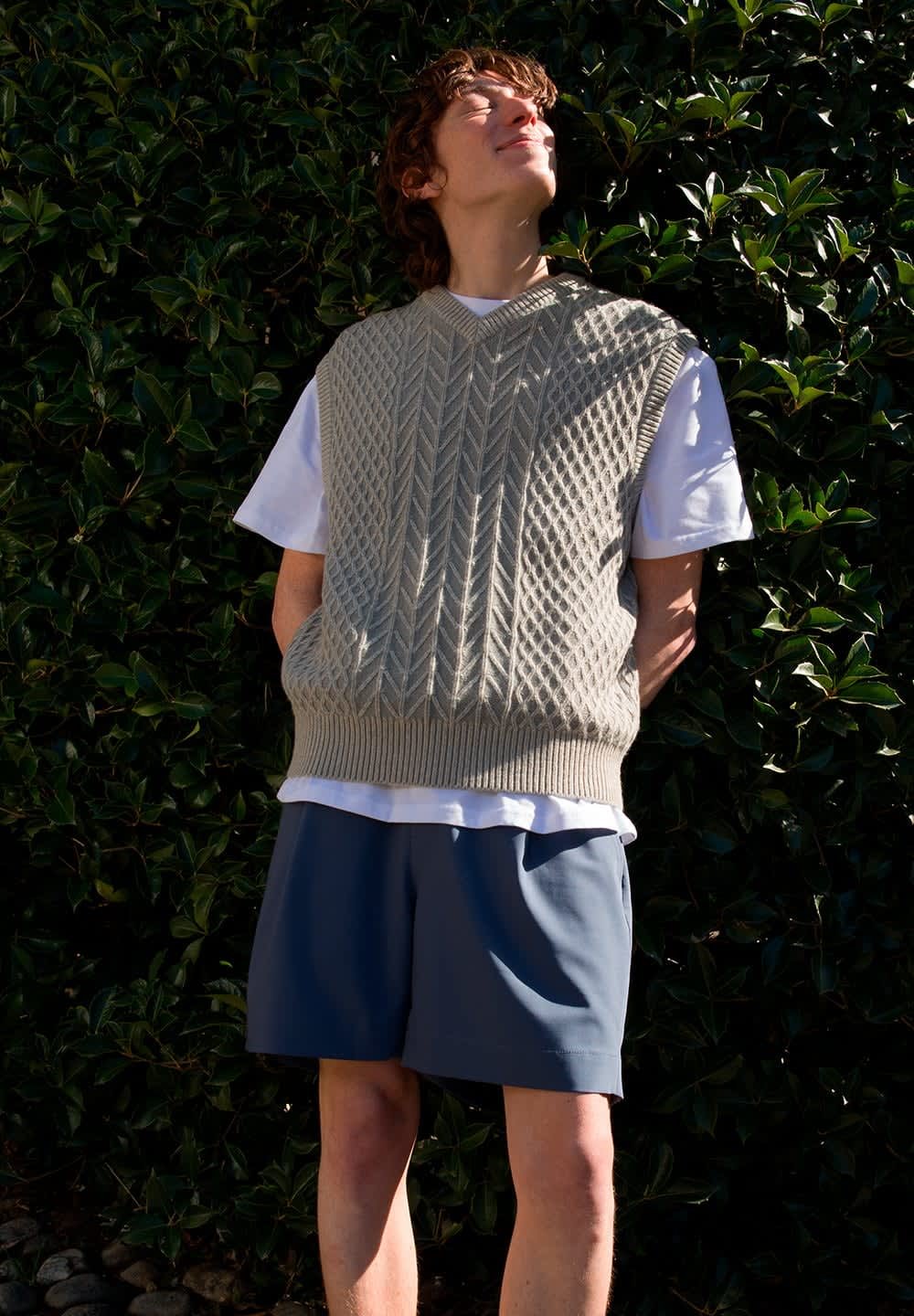 Maglione senza maniche lavorato a maglia beige con un motivo a diamante sopra una t-shirt bianca, abbinato a pantaloni corti blu, il tutto di fronte a uno sfondo di arbusti verdi.