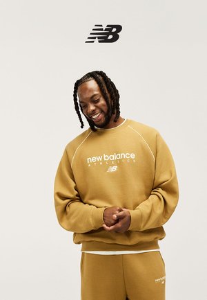 Man met gevlochten haar die lacht, gekleed in een mosterdgele New Balance Athletics trui en bijpassende broek tegen een effen achtergrond.