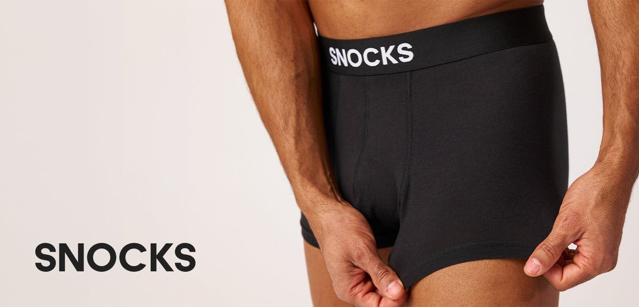 Man draagt zwarte Snocks boxershorts en past het been aan tegen een neutrale beige achtergrond.
