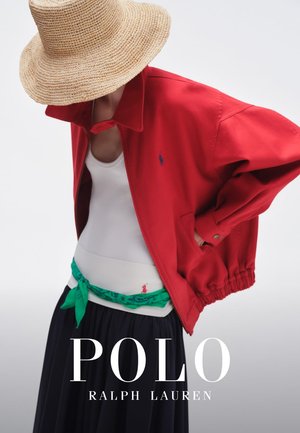 Persona con un sombrero de paja de ala ancha, chaqueta roja, camiseta blanca sin mangas, cinturón bufanda verde y falda oscura plisada, posando con la cabeza inclinada hacia abajo.