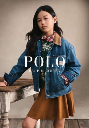 Joven girl apoyada en una mesa rústica de madera, vistiendo una chaqueta de mezclilla sobre un suéter estampado, camisa a cuadros y falda marrón contra un fondo liso.