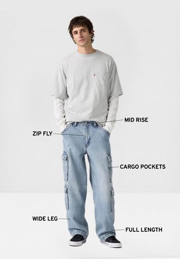 Joven con camiseta de manga larga gris claro superpuesta y vaqueros cargo de pierna ancha y largo completo azul claro con cremallera y talle medio.