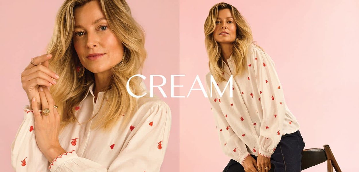 Blonde Frau in einer cremefarbenen Bluse mit roten Kirschmotiven posiert vor einem blassrosa Hintergrund, sitzt und steht selbstbewusst.