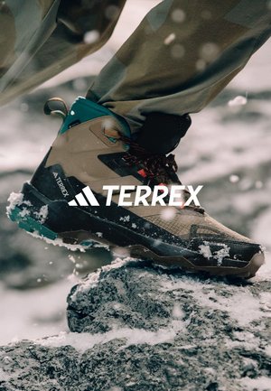 Stivale da trekking impermeabile marrone e teal con superficie testurizzata, dotato di suola gommata e lacci rossi, posizionato su terreno roccioso.