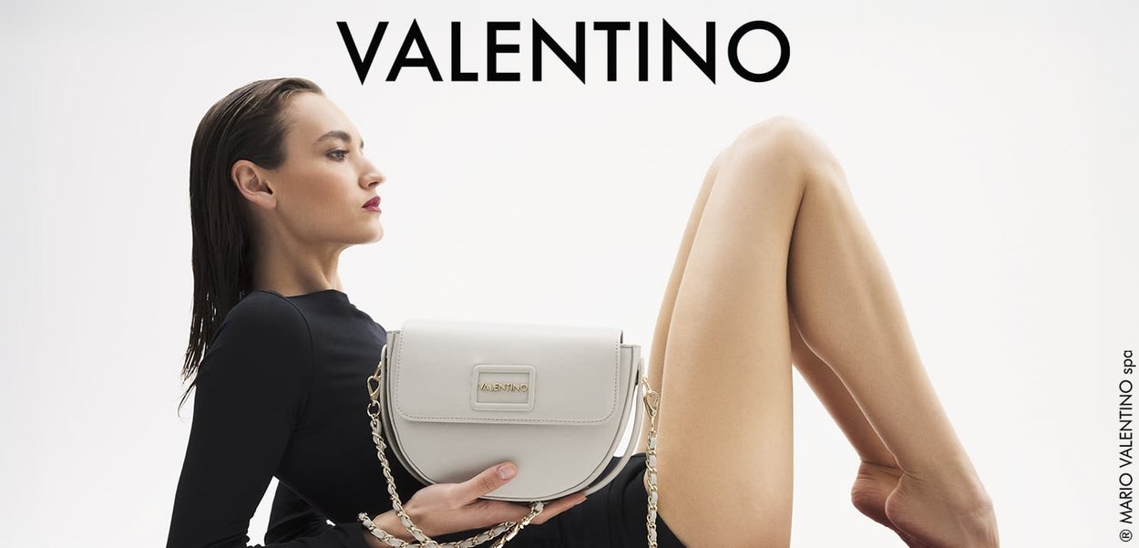 Torebka Valentino w off-white skórze, o strukturalnym kształcie, ze złotymi akcentami i łańcuszkowym paskiem. Modelka pozuje w czarnym, długim rękawie.