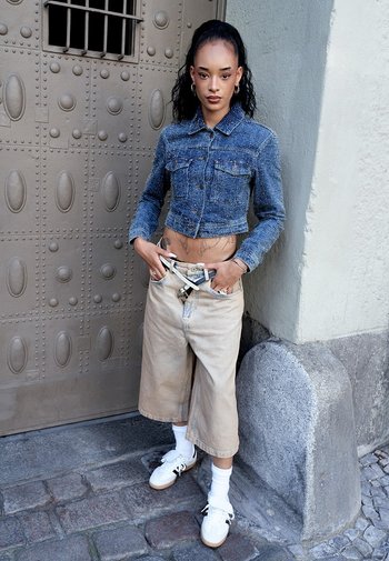 Denim cropped jacket, lichtbeige wijde broek, witte sneakers en een gestreepte riem. Stedelijke omgeving met een textuurrijke metalen deur op de achtergrond.