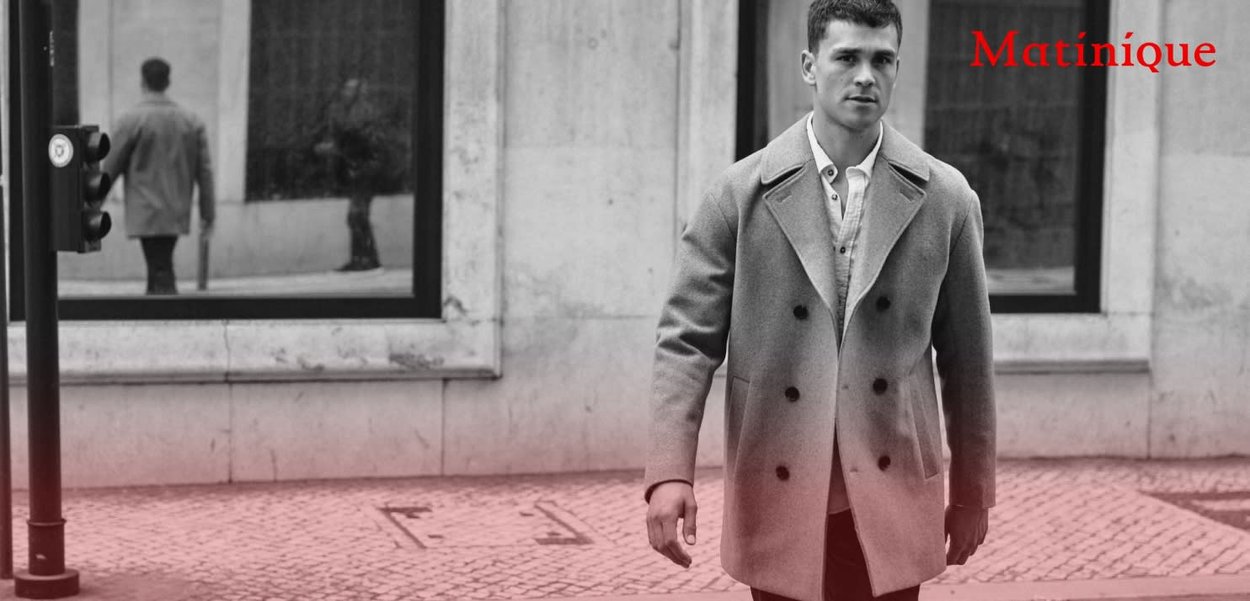 Grauer Doppelreiher-Mantel mit großen Revers, getragen über einem hellen Hemd. Das Modell steht auf einer Straße mit Spiegelungen in Glasfenstern, in der Nähe einer Ampel.