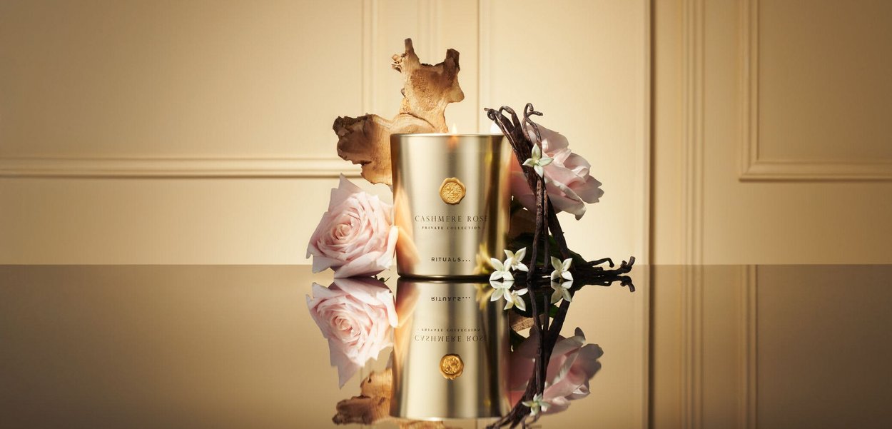 Candela d'oro accesa etichettata "Cashmere Rose" circondata da rose rosa, baccelli di vaniglia, fiori bianchi e un pezzo di legno su una superficie riflettente.