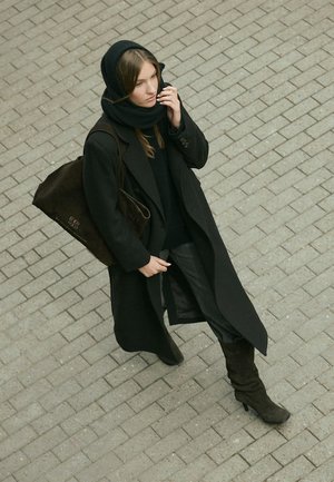 Lange zwarte jas en zwarte sjaal gedragen over een zwarte trui en zwarte broek. Bruine schoudertas met logo. Donkere kniehoge laarzen. Geplaveide wandelweg.