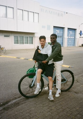 Twee personen op een lichtblauwe fiets met een groene tas. De grond is bestraat en er zijn industriële gebouwen op de achtergrond.