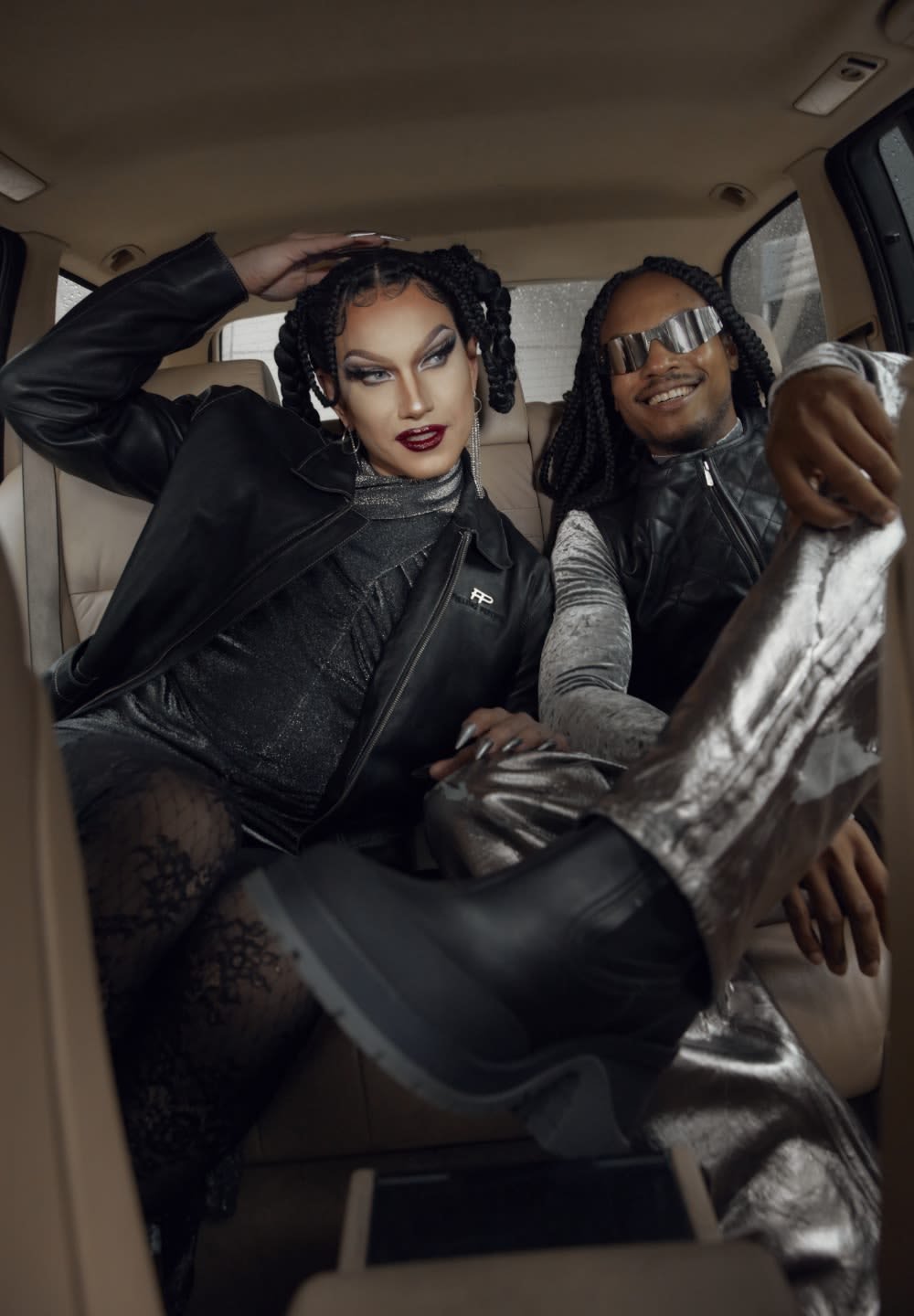 Twee personen in een auto. De ene draagt een zwarte leren jas en zilveren leggings, terwijl de ander een glanzend zwart vest en metalen broek heeft.