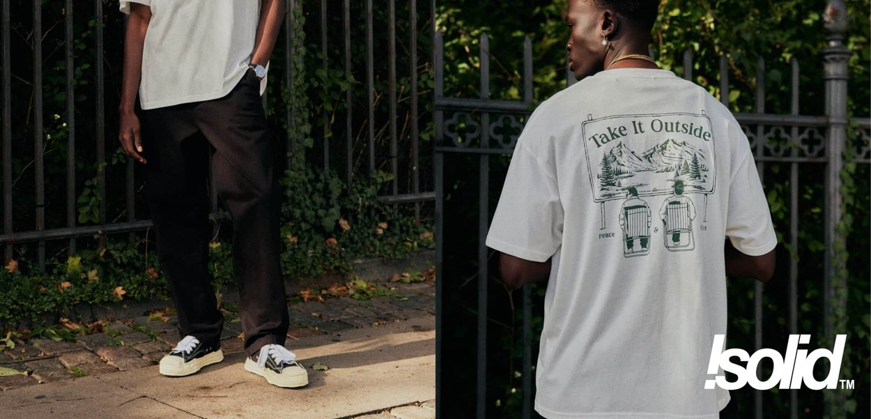 Mann trägt weißes T-Shirt mit Schriftzug "Take It Outside" und Berggrafik, steht draußen auf einem gepflasterten Weg in der Nähe eines schwarzen Eisenzauns.