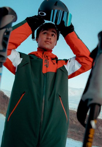 Ski jakke i grøn med orange og hvide detaljer, der har en høj krave, lynlåslommer, og en person der holder en sort hjelm.