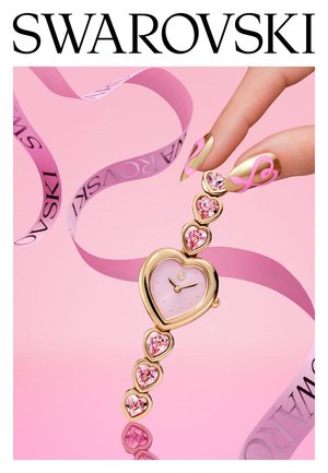Gouden horloge in de vorm van een hart met roze edelstenen, vastgehouden door een hand met gouden en roze nagels met hartmotief, op een roze achtergrond met een Swarovski-lint.