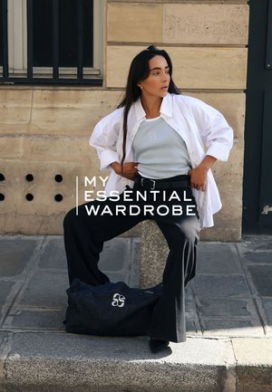 Pantalons larges noirs, débardeur bleu clair et chemise oversize blanche. Sac gris avec logo posé à côté. Fond en pierres pavées.