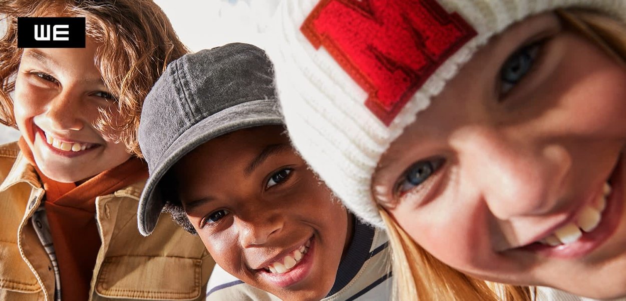 Drei Kinder lächeln, tragen eine Jeansjacke, eine graue Kappe und eine weiße Strickmütze mit einem roten "M"-Detail. Helles Licht hebt ihre Mienen hervor.