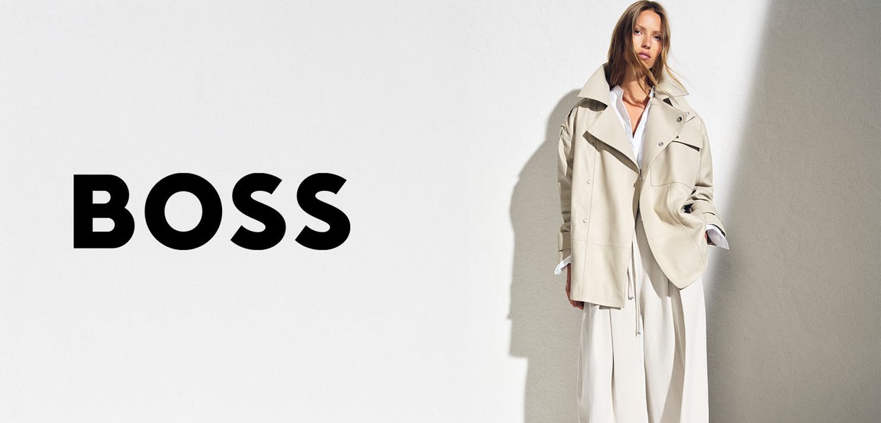 Frau trägt eine beige übergroße Jacke und ein weißes Outfit und steht vor einem neutralen Hintergrund, links davon in kräftigem Schwarz der Schriftzug "BOSS".