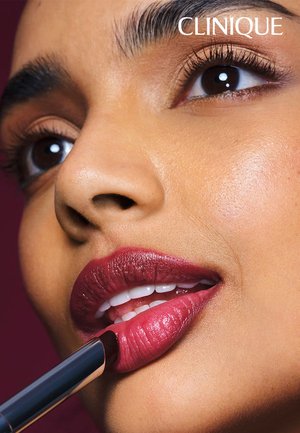 Primo piano di una donna che si applica un rossetto rosso scuro con un logo clinico sopra il suo occhio destro su uno sfondo scuro.