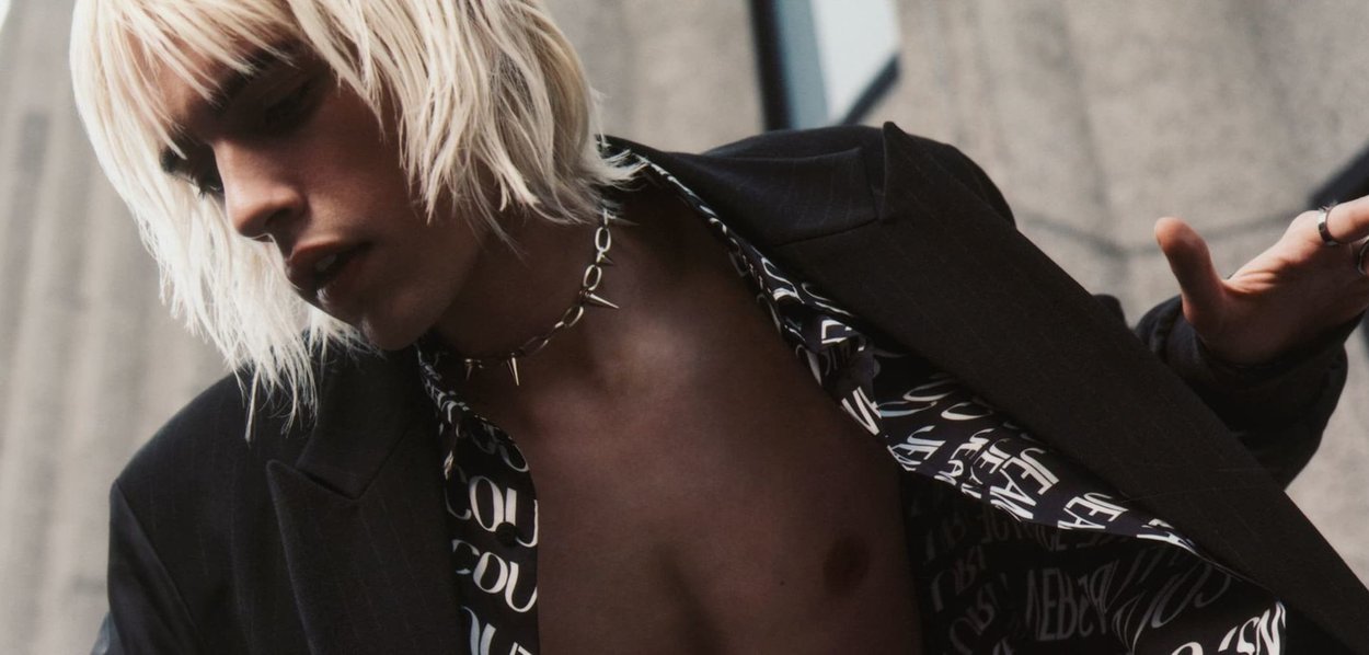 Blazer noir sur une chemise à motifs avec un texte blanc, avec un collier à pointes en argent, gros plan sur une personne aux cheveux blonds clairs.