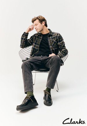 Schwarze Lederschuhe mit einer dicken Gummisohle, getragen mit dunklen Jeans und einem karierten Hemd über einem schwarzen T-Shirt. Modell sitzt auf einem Drahtstuhl.