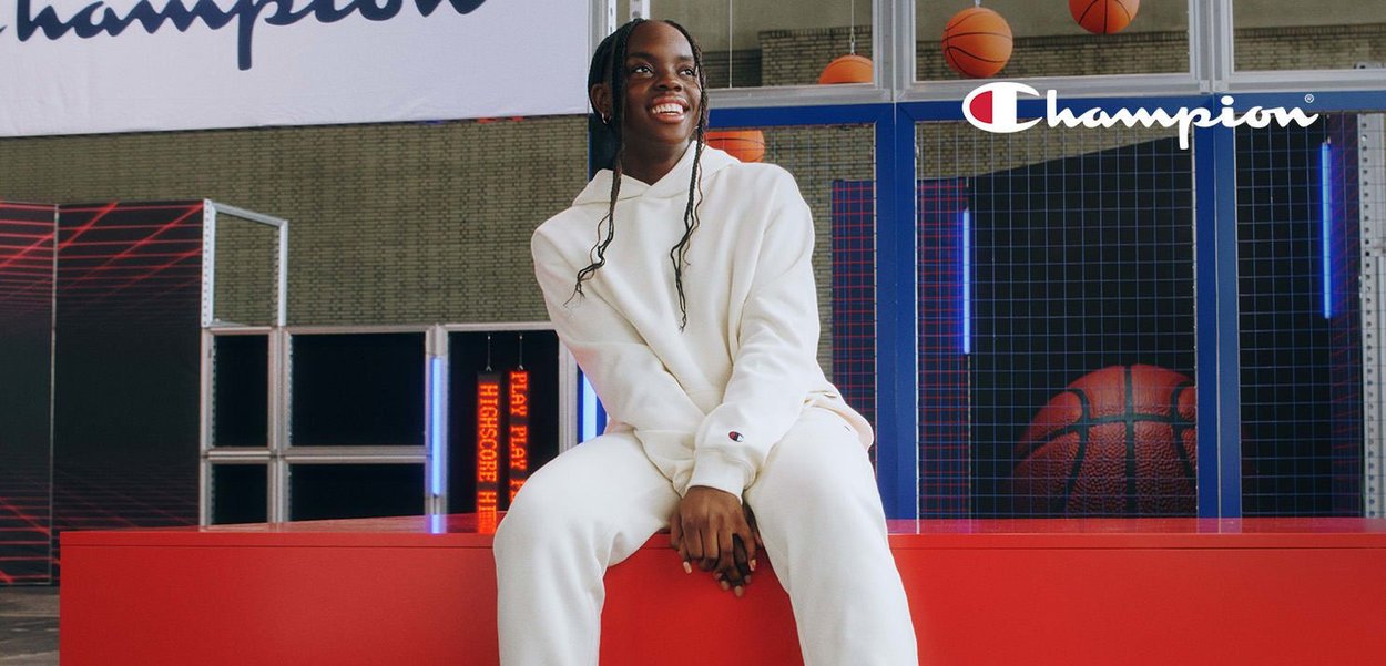 Witte hoodie en broek van Champion, met logodetail. Helderrode displayplatform. De achtergrond heeft neonlichten en basketbal. 