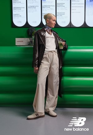 Manteau en cuir marron porté sur un t-shirt blanc avec l'inscription « new balance », pantalon beige à jambes larges, et baskets beige avec des accents gris. Fond vert.