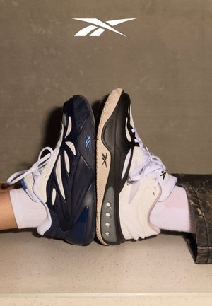 Two pairs of Reebok sneakers in black, white, and blue colors with white socks, soles touching, against a neutral brown background.