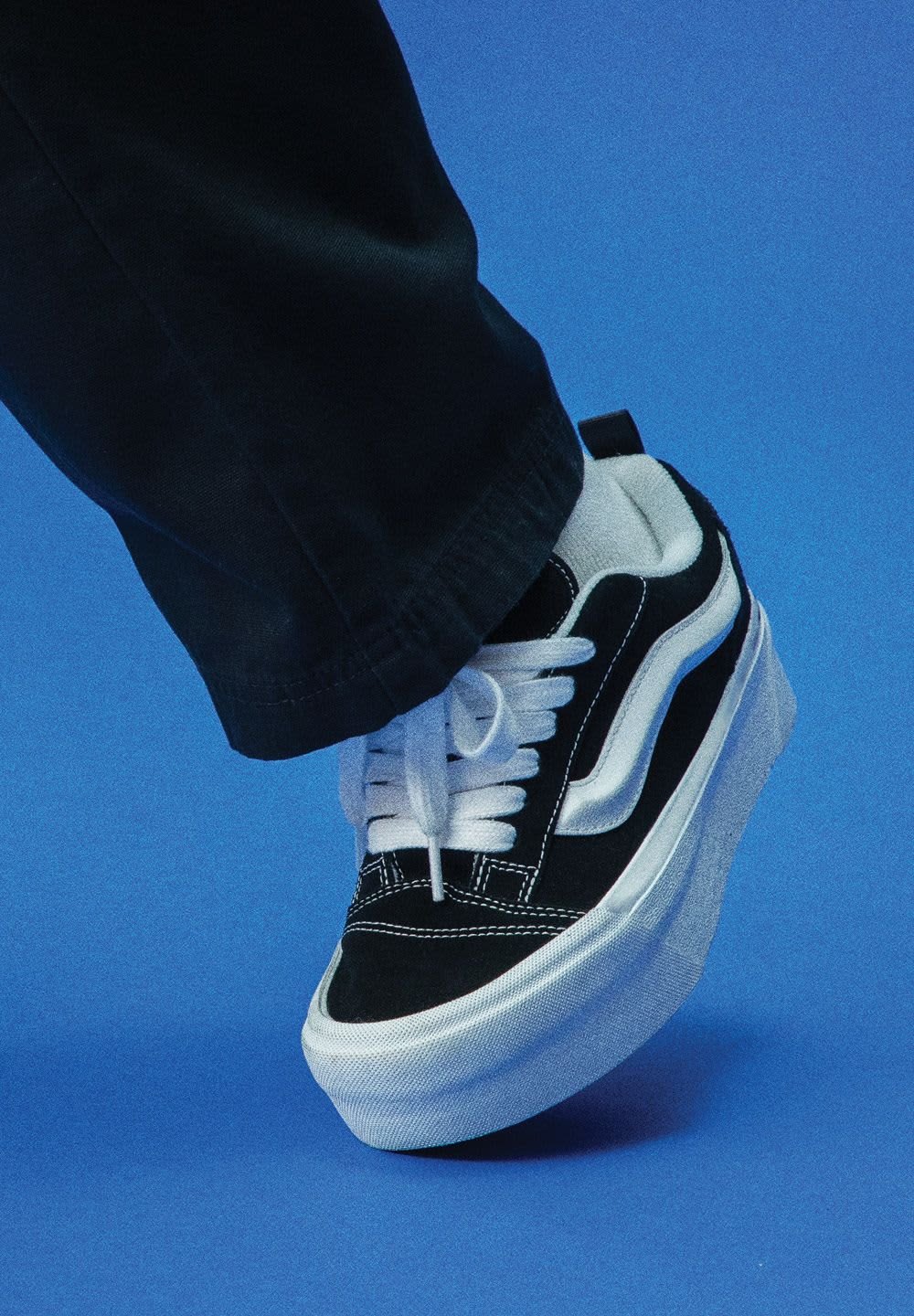 Schwarze und weiße Low-Top Sneakers mit einem Obermaterial aus Wildleder, weißer Schnürung und einer dicken weißen Gummisohle vor einem blauen Hintergrund.