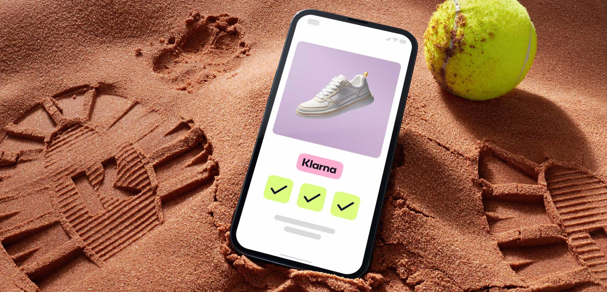 Un smartphone sur le sable affiche une chaussure de sport blanche avec une semelle texturée sur un fond violet pastel, à côté d'une balle de tennis.