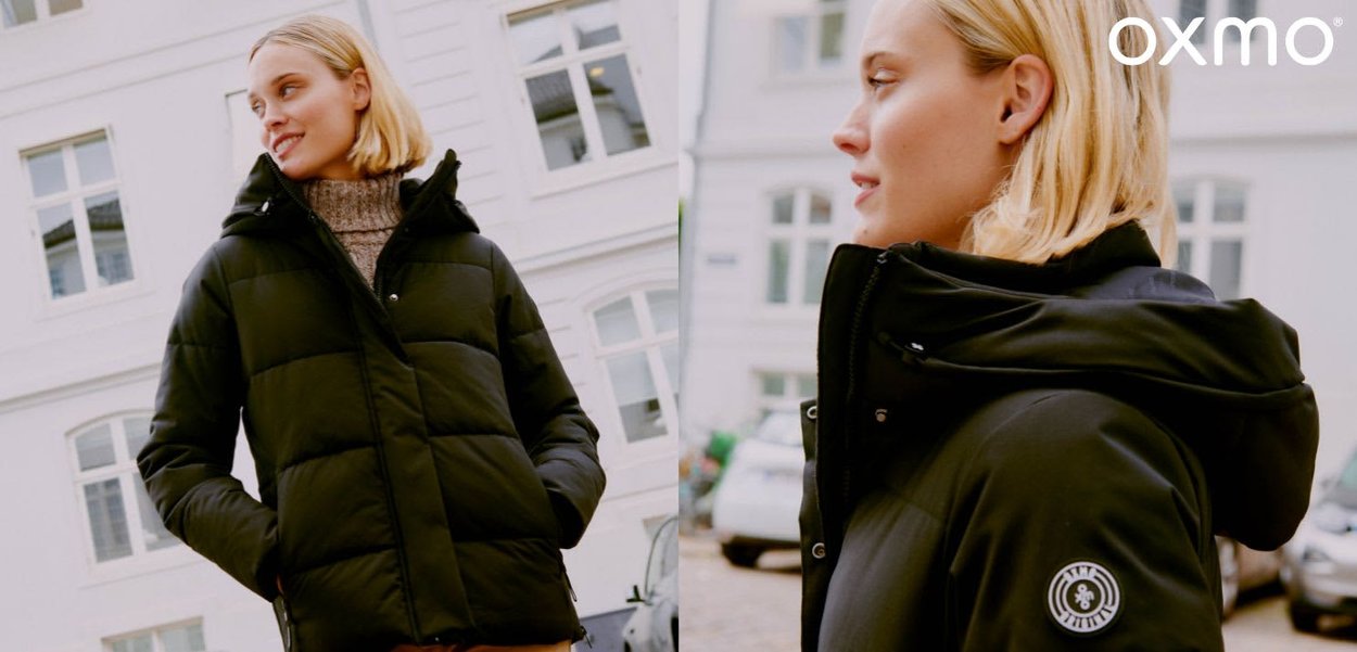 Femme blonde portant une doudoune noire et un pull col roulé marron en plein air dans un cadre urbain avec des bâtiments blancs.