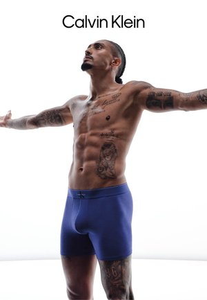 Uomo senza maglietta con tatuaggi che allarga le braccia, indossando boxer Calvin Klein blu, volto leggermente rivolto verso l'alto su sfondo bianco.