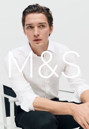 Young man with styled brown hair wearing a white button-up shirt and black pants, seated on a white chair against a plain background.
