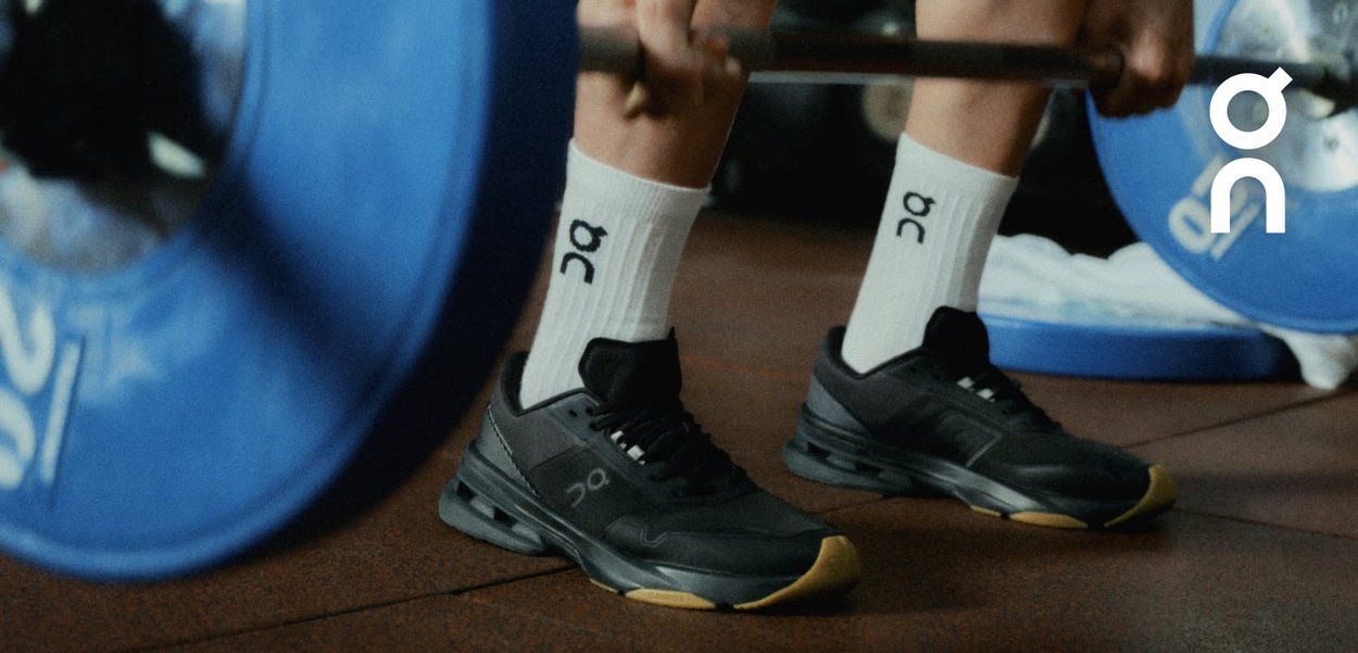 Black athletic shoes with a cushioned sole and a gold accent, paired with white socks displaying a logo, on a gym floor near weight plates.