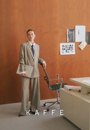 Woman in beige suit holding a laptop, standing by a black office chair in a minimalist workspace with design sketches on a wood wall.