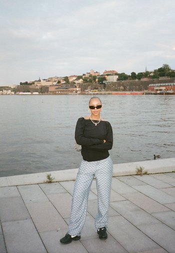 Zwarte lange mouwen top, gemêleerde lichtblauwe broek, zwarte schoenen. Accessoires omvatten zonnebril en gelaagde kettingen. Water en gebouwen op de achtergrond.