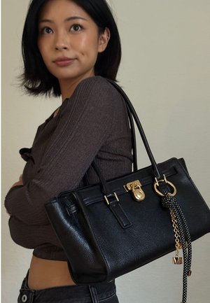 Junge Frau in einem dunklen Langarmoberteil, die eine schwarze Lederhandtasche mit goldenen Akzenten und einem dekorativen schwarz-weißen Quasten trägt.