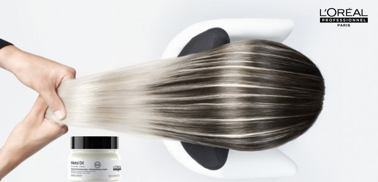 Mains tenant des cheveux longs, lisses et brillants avec des racines foncées et des pointes platine au-dessus d'un bassin blanc, avec un produit L’Oréal Metal DX à proximité.