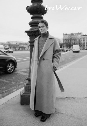 Longue veste grise en mélange de laine avec un col structuré et des poches latérales, portée sur un pull en tricot, associée à un pantalon à jambes larges et des chaussures à talons blocs.