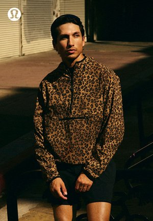 Hombre con chaqueta tipo jersey de estampado de leopardo y pantalones cortos negros, de pie al aire libre con poca luz cerca de un edificio con persianas cerradas.