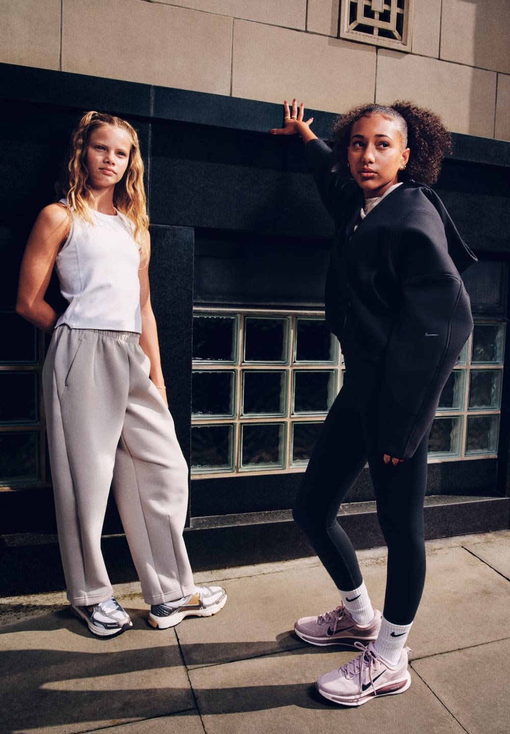 To unge kvinder står op ad en væg af sorte og glasblokke iført afslappede sporty outfits og sneakers udendørs på en solrig dag.