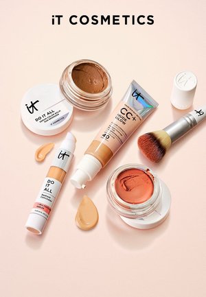 iT Cosmetics Bronzer, Concealer, CC+ Nude Glow Tube, Rouge in Tiegeln, Make-up-Pinsel und Produktproben auf blassrosa Hintergrund.